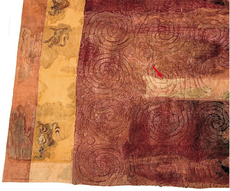Шерстяной ковер, простеганный спиральным орнаментом, из погребальной камеры 20-го Ноин-Улинского кургана. На покрывающей ковер ткани – светлый зубчатый орнамент (мерлон) – показан красной стрелкой