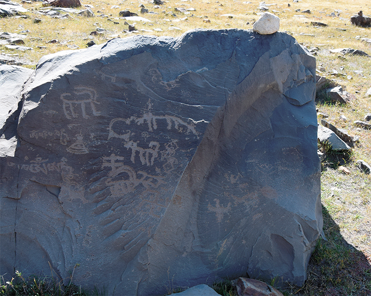 Akshow petroglyphs. A composition imbibed with Buddhist symbolism, consisting of ibex images created in all the known styles (silhouette bitriangular, geometric, and linear), alternating with images of right and left swastikas, a small stupa, a shell, and an ascetic with a skull-like head and arms crossed and raised upwards. His body is overlapped by an image of a small animal resembling a dog, created with the same thin metal instrument as both the man himself and the shell. The surface of this stone also bears a double inscription of the Buddhist mantra Om mani padme hum and, below, the mantra Om a hum. In the Chinese Buddhist tradition, this mantra was called Sanzi zongchi zhou, or “Mantra [in] three words [that grants] absolute control [over passions]” (read and interpreted by S.Kh. Shomakhmadov, Cand. Sci. (History), a senior researcher at the Institute of Oriental Manuscripts, Russian Academy of Sciences, for which the author of this article is immensely grateful to him). All these images are interconnected and date back to the period after the “second advent of Buddhism” in Ladakh in the 8th century. Another facet of the stone shows an image a swastika-like bird (bottom left). Zanskar, 2019