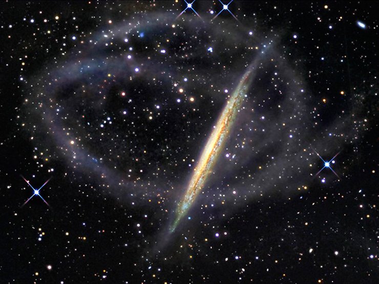 Светлые кольца вокруг галактики – это не дефект фотографии, а поток звёзд, в который превратился один из спутников этой галактики. Поначалу это было небольшое самостоятельное звёздное скопление, но приливом его вытянуло вдоль орбиты и размазало по ней