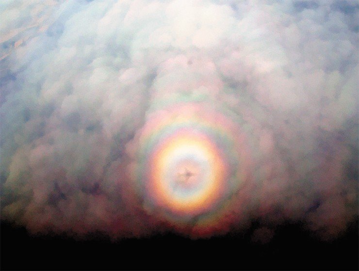 Тень самолета на облаках и окружающий ее ореол, который вызван дифракцией света на крупном объекте. Происхождение цветных колец имеет другую природу. Контраст деталей подчеркнут при обработке снимка. Снимок сделан с меньшей высоты, чем фото выше
